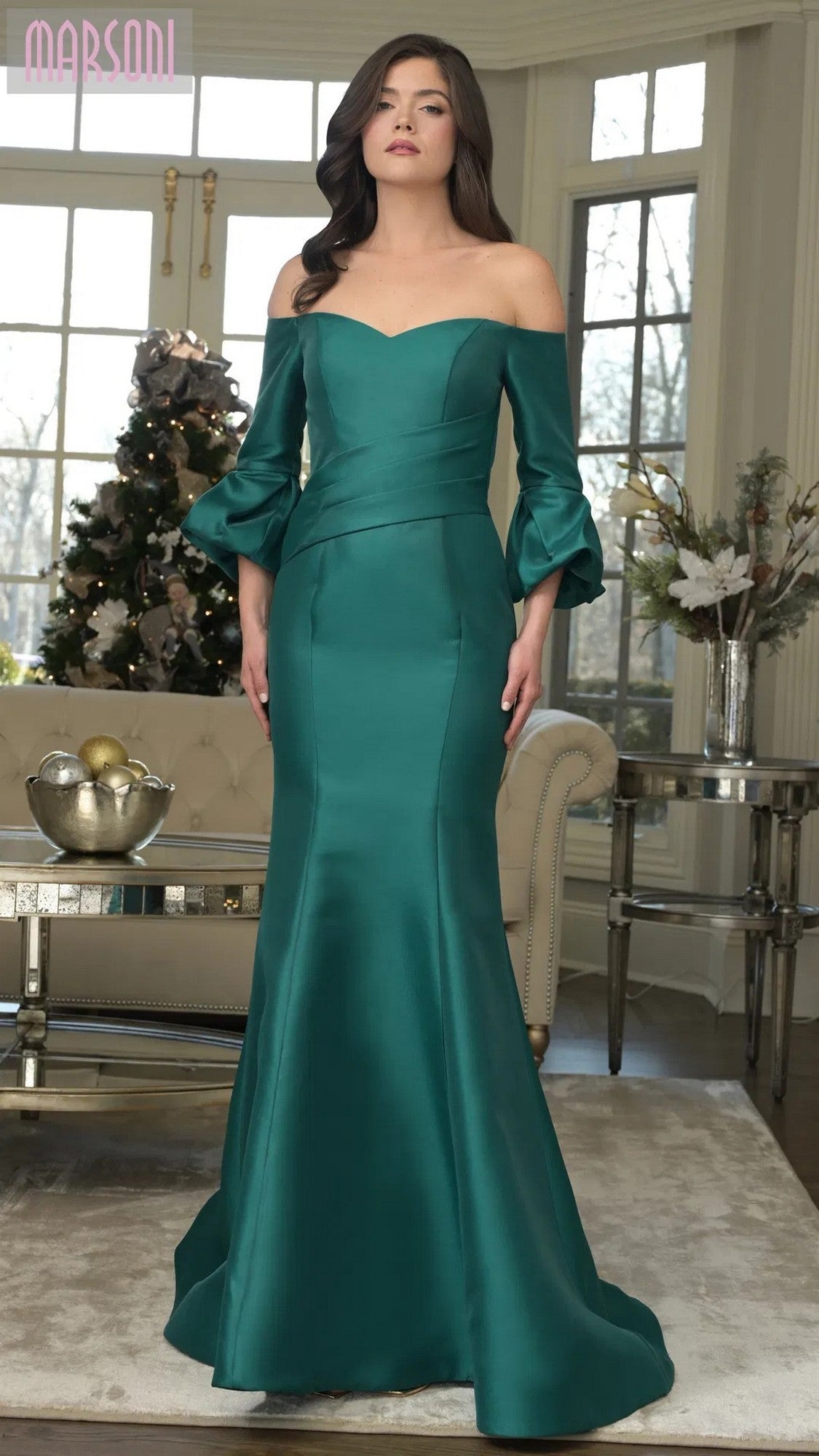 A woman poses indoors in the Marsoni MV1355, an emerald long formal dress with off-the-shoulder style, mermaid silhouette, and flared sleeves. A decorated Christmas tree enhances the festive scene.